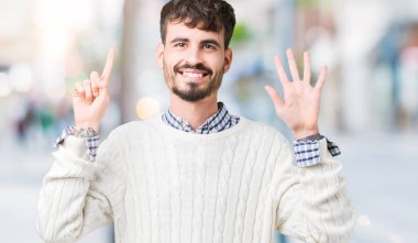 Gösterilen arka plan üzerinde kış kazak giyen genç yakışıklı adam izole ve parmakları ile işaret kendinden emin ve mutlu gülümseyerek süre 6 numara.