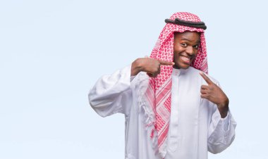 Geleneksel keffiyeh kendine güvenen gösterilen ve işaret parmakları diş ve ağız ile gülümseyen izole arka plan üzerinde giyen genç Arapça Afrika adam. Sağlık kavramı.