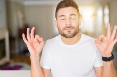 Casual beyaz t-shirt evde dinlenmek giydiği ve gözleri ile gülümseyen yakışıklı delikanlı parmak ile meditasyon jest yapıyor kapalı. Yoga kavramı.