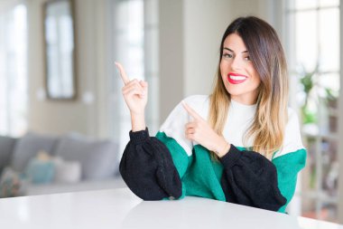 Evde kış kazak giyen genç güzel kadın gülümseyerek ve iki elleri ve parmakları ile tarafına işaret kamera bakıyor.