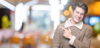 Yakışıklı delikanlı izole arka neşeli bir gülümseme ile yüz el ve parmak kadar mutlu ve doğal ifade yüzünde yanında işaret içinde kış palto giyiyor