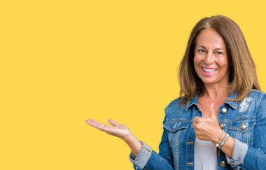 Üzerinde rahat denim ceket giyen güzel orta yaş kadın arka plan gösteren palm el ve iş tamam jestle başparmak yukarıya, mutlu ve neşeli gülümseyen izole