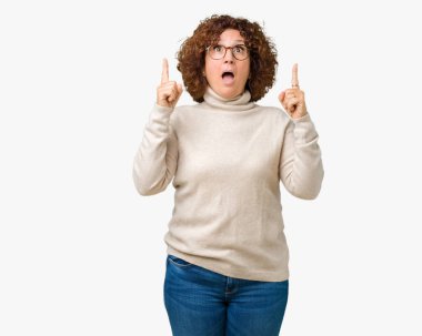 Boğazlı kazak ve gözlük izole arka plan üzerinde giyen güzel orta ager üst düzey kadın hayrete ve yukarıya bakıp parmakları ile işaret şaşırttı ve silah kaldırdı.