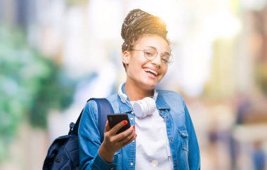 Genç duran ve diş gösterilen kendine güvenen bir gülümsemeyle gülümseyerek mutlu bir yüz ile izole arka plan üzerinde akıllı telefon kullanan saç Afro-Amerikan öğrenci kız örgülü