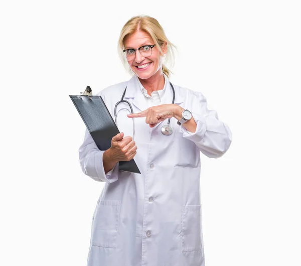 中年金发碧眼的医生妇女拿着剪贴板在孤立的背景非常高兴用手和手指指着 — 图库照片