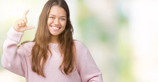 孤立した背景の笑顔と自信を持ってカメラを見ながら指でサイズ記号をやって手でジェスチャーをピンク冬のセーターを着た若い美しいブルネット女性 測定概念 — ストック写真
