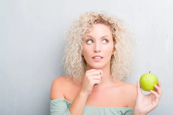 年轻的金发碧眼的女人在粗野的灰色背景下吃绿色苹果严肃的脸思考问题 很困惑的想法 — 图库照片