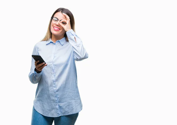年轻美丽的女商人短信使用智能手机在孤立的背景与快乐的脸微笑做确定的标志与眼睛看着手指 — 图库照片