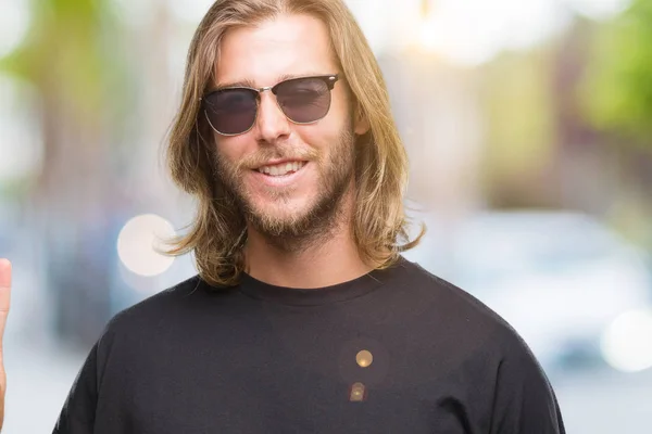 Young handsome man with long hair wearing sunglasses over isolated background smiling with happy face winking at the camera doing victory sign. Number two.