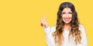 Mutlu yüz zafer işareti yaparken kameraya göz kırpıyor ile gülümseyen beyaz kazak giyen genç güzel kadın. İki numaralı.