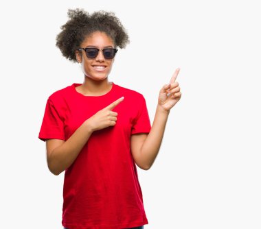 Genç afro Amerikalı kadın gülümseyerek ve iki elleri ve parmakları ile tarafına işaret kamera bakarak izole arka plan üzerinde güneş gözlüğü takıyor.