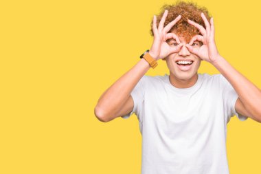 Dilini, parmaklarının arasından baktığını yapışmasını dürbün gibi ok jest yapıyor casual beyaz t-shirt giyen afro saçlı Genç yakışıklı adam. Deli ifade.