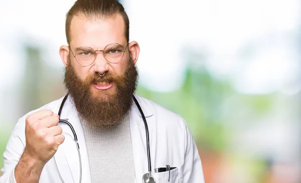 Young blond doctor man with beard wearing medical coat angry and mad raising fist frustrated and furious while shouting with anger. Rage and aggressive concept.