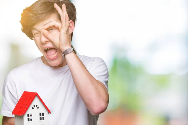 Young man holding house over isolated background with happy face smiling doing ok sign with hand on eye looking through fingers