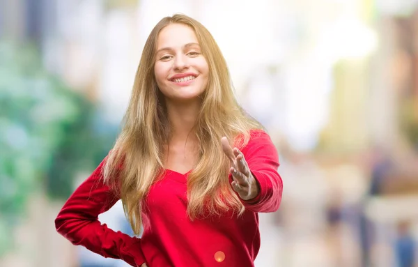 若い美しい金髪女性の挨拶と歓迎フレンドリーな提供ハンドシェイク笑みを浮かべて分離の背景に赤いセーターを着ています ビジネスの成功 — ストック写真