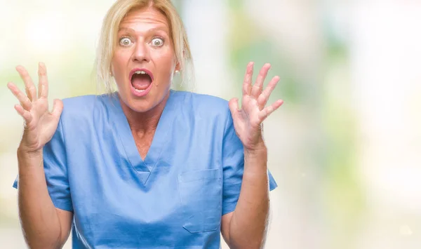 中年金发碧眼的女人穿着医生护士制服在孤立的背景疯狂和疯狂的喊声和大喊大叫与侵略性的表情和手臂提高 挫折的概念 — 图库照片