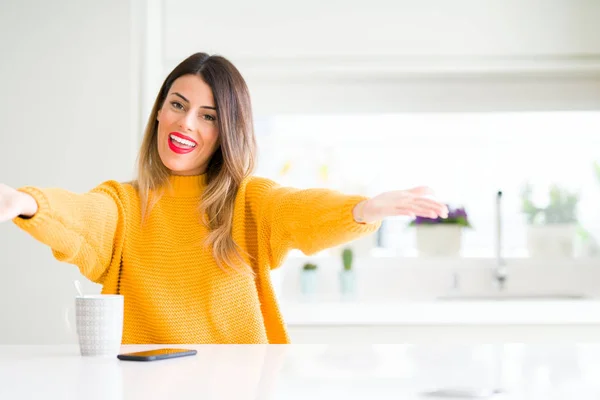 Young beautiful woman drinking a cup of coffee at home looking at the camera smiling with open arms for hug. Cheerful expression embracing happiness.