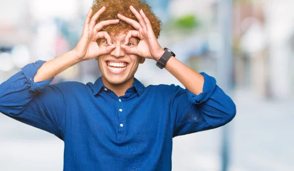 Young handsome elegant man with afro hair doing ok gesture like binoculars sticking tongue out, eyes looking through fingers. Crazy expression.
