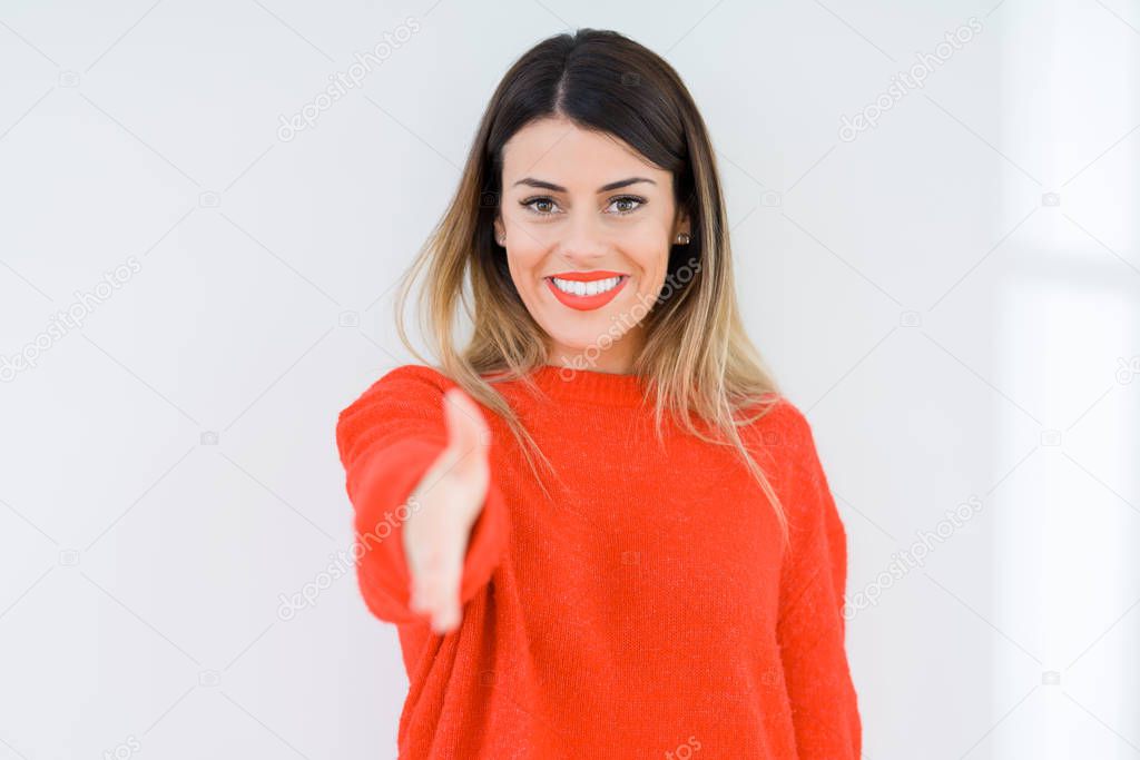 Young woman wearing casual red sweater over isolated background smiling friendly offering handshake as greeting and welcoming. Successful business.