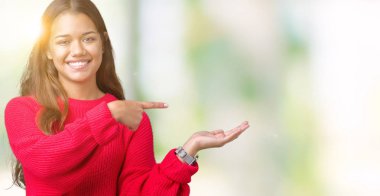 Hayret ve el ile sunulması ve parmak ile işaret ederken kameraya gülümseyen izole arka plan üzerinde kırmızı kış kazak giyen genç güzel esmer kadın.