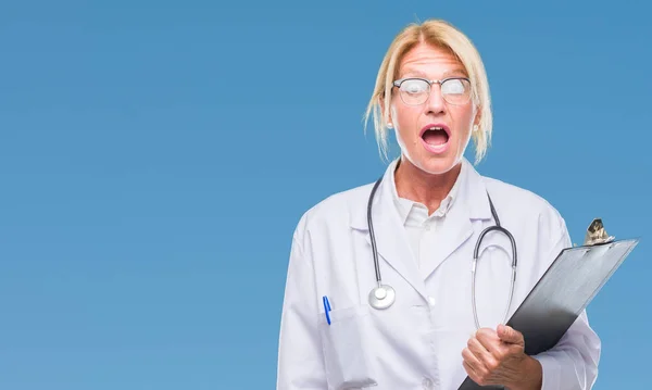 Middle age blonde doctor woman holding clipboard over isolated background scared in shock with a surprise face, afraid and excited with fear expression