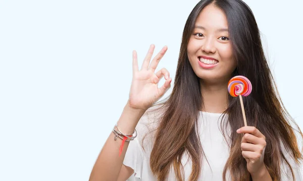 優秀なシンボルの指で サインをして孤立した背景に棒付きキャンディーを食べる若いアジア女性 — ストック写真
