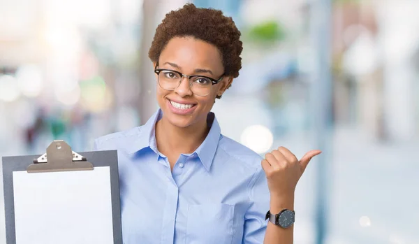 指していると幸せそうな顔を笑顔で側まで親指で示す分離の背景にクリップボードを保持している若いアフリカ系アメリカ人ビジネス女性 — ストック写真