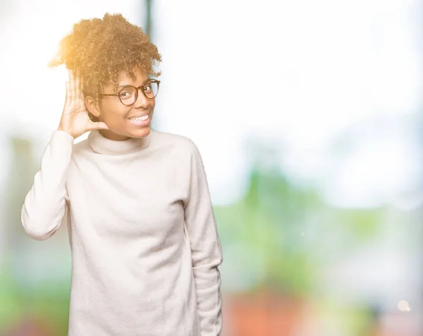 Beautiful young african american woman wearing glasses over isolated background smiling with hand over ear listening an hearing to rumor or gossip. Deafness concept.