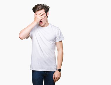 Mahcup ifadeyle parmaklarımın arasından seyir yüz ve gözler el ile kapsayan şok bakmak izole arka plan üzerinde rahat beyaz t-shirt giyen genç yakışıklı adam.