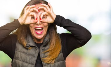 Dilini, parmaklarının arasından baktığını yapışmasını dürbün gibi ok jest yapıyor kış yelek giyen güzel orta yaş kadın. Deli ifade.