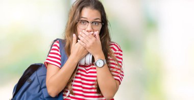 Üzerinde kulaklık ve sırt çantası giyen genç güzel esmer öğrenci kadın arka plan ağız hata için elleriyle kapsayan şok izole. Gizli kavramı.