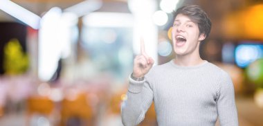 Genç yakışıklı adam başarılı fikirle izole arka plan işaret parmağı üzerinde kış kazak giyiyor. Çıktıktan ve mutlu. Bir numara.