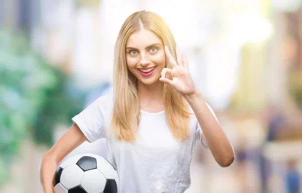 優秀なシンボルの指で サインをして孤立した背景にサッカー ボールを保持している若い美しいブロンドの女性 — ストック写真