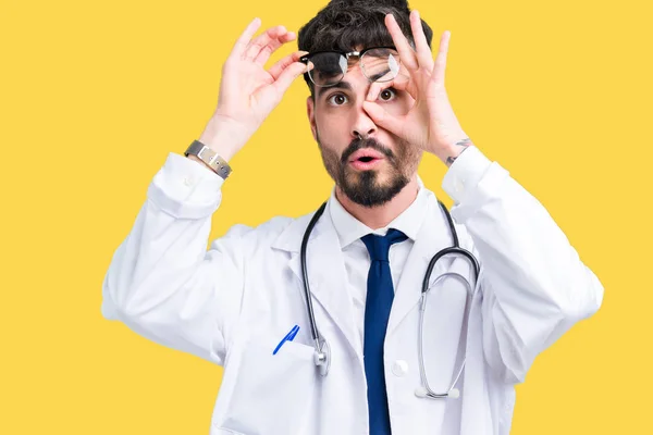 Young doctor man wearing hospital coat over isolated background doing ok gesture shocked with surprised face, eye looking through fingers. Unbelieving expression.