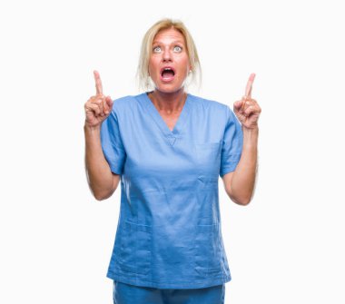 İzole arka plan üzerinde doktor hemşire üniforması giyen orta yaş sarışın kadın hayrete ve yukarıya bakıyor ve parmakları ile işaret ve silah kaldırdı şaşırttı.