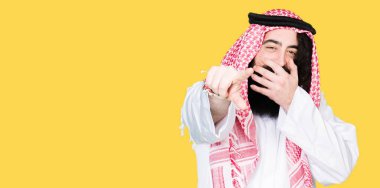 Arap iş adamı geleneksel keffiyeh giymiş uzun saçlı eşarp gülme sizin parmak el kamerasıyla ağzına, utanç ifadesi işaret eden