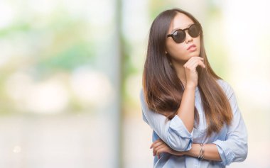 Genç Asyalı kadın elle çene düşünme hakkında soru, dalgın ifade izole arka plan üzerinde güneş gözlüğü takıyor. Düşünceli yüzüyle gülümsüyor. Şüphe kavramı.