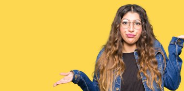 Gözlük ve denim ceket clueless ve şaşkın bir ifadeyle kollar ve eller kaldırdı giyen genç güzel kadın. Şüphe kavramı.