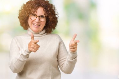 Boğazlı kazak ve gözlük için fotoğraf makinesi ile mutlu ve komik yüz parmakları işaret izole arka plan üzerinde giyen güzel orta ager üst düzey kadın. İyi enerji ve vibes.