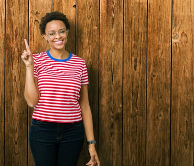 Güzel genç Afrikalı-Amerikalı kadın ve kendinden emin ve mutlu gülümseyerek süre parmak numaralı yukarıyı gösteren izole arka plan üzerinde gözlük.