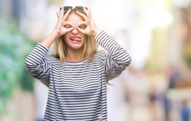 Genç güzel sarışın kadın dilini, parmaklarının arasından baktığını yapışmasını dürbün gibi ok jest yapıyor izole arka plan üzerinde güneş gözlüğü takıyor. Deli ifade.