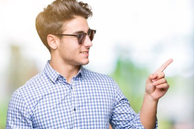 Kameraya bakarak yan el ve parmak ile işaret eden genç yakışıklı adam yüzünde kocaman bir gülümseme ile izole arka plan üzerinde güneş gözlüğü takıyor.