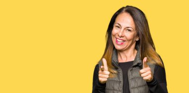 Kış yelek işaret parmakları için fotoğraf makinesi ile mutlu ve komik yüz giyen güzel orta yaş kadın. İyi enerji ve vibes.