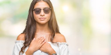 Kapalı gözler ve yüzünde minnettar jest ile göğüs üzerinde eller genç güzel Arap kadın ile gülümseyen izole arka plan üzerinde güneş gözlüğü takıyor. Sağlık kavramı.