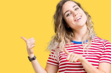 İzole arka plan üzerinde sarışın güzel genç kadın kendine güvenen gülümseyerek arkasından el ve başparmak yukarıya, işaret