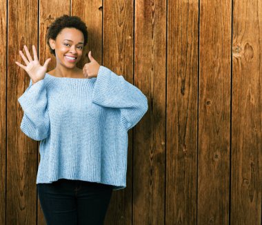 Gösterilen ve parmakları ile işaret izole arka plan üzerinde bir kazak giyen genç güzel Afrikalı-Amerikalı kadın kendinden emin ve mutlu gülümseyerek süre 6 numara.