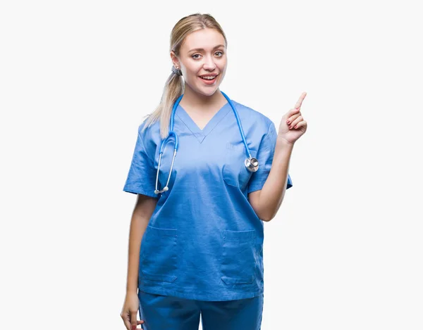 若い金髪の外科医医師女性の顔に大きな笑みを浮かべて隔離された背景にカメラを見ている側の手と指で指す — ストック写真