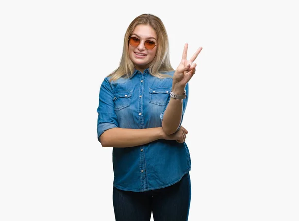 若い白人女性の勝利のサインを行うカメラでウィンクしている幸せそうな顔を浮かべて孤立の背景の上にサングラスをかけています — ストック写真