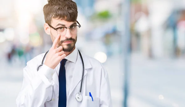 Young doctor man wearing hospital coat over isolated background Pointing to the eye watching you gesture, suspicious expression