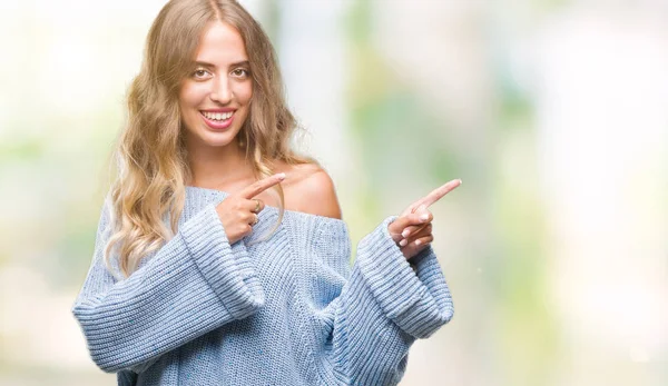 Gülümseyen ve iki elleri ve parmakları ile tarafına işaret kamera bakarak izole arka plan üzerinde kış kazak giyen güzel genç sarışın kadın.